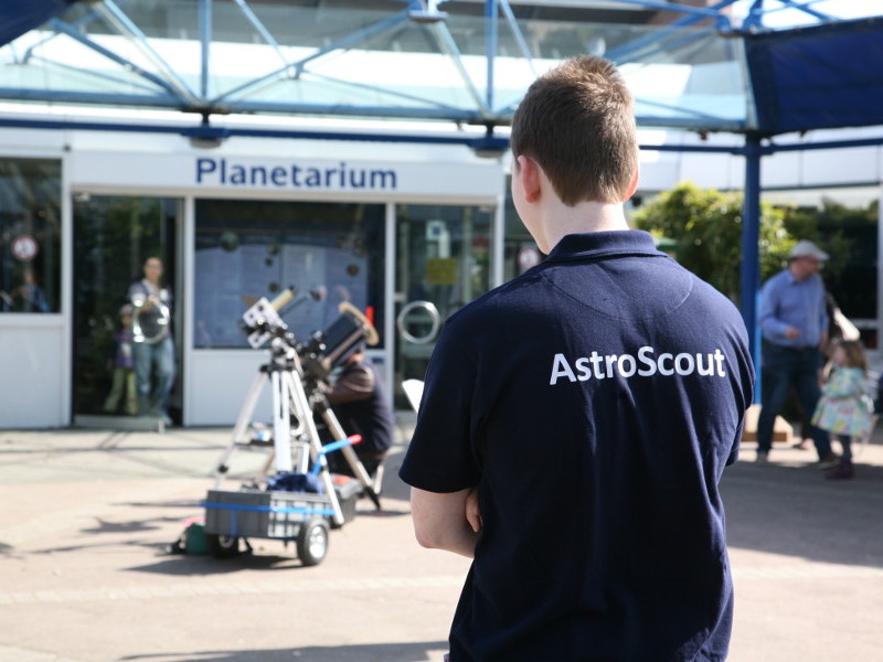 Link zu Informationen über das Mentorenprojekt „AstroScout“ auf der Homepage des Planetarium Mannheim - Das Bild zeigt einen Jugendlichen mit dunkelblauem T-Shirt und der weißen Aufschrift „AstroScout“ von hinten vor dem Eingangsbereich des Planetariums.