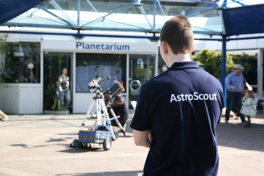Das Bild zeigt den Oberkörper eines Jugendlichen von hinten vor dem Eingangsbereich des Planetarium Mannheim. Er trägt ein dunkelblaues T-Shirt mit der weißen Aufschrift „AstroScout“. Zwischen ihm und dem Planetarium stehen zwei Teleskope.