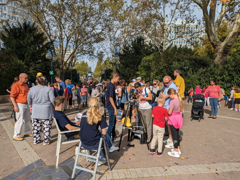Link zu Informationen über Aktionen der AstroScouts auf der Homepage des Planetarium Mannheim – Das Bild zeigt Menschen auf dem Vorplatz des Planetariums beim Beobachten der Sonne unter Anleitung der AstroScouts des Planetariums.