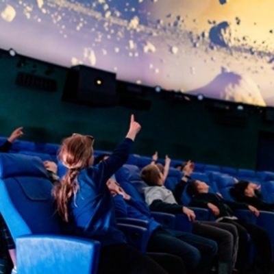Link zum Bereich „Bildung“ auf der Homepage des Planetarium Mannheim - Das Bild zeigt eine Gruppe von Kindern im Kuppelsaal des Planetariums. Sie sehen sich eine Planetariumsshow an. Mehrere Kinder zeigen mit ausgestreckten Armen nach oben.