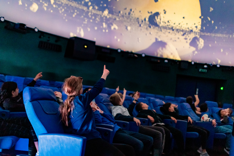 Das Bild zeigt eine Gruppe von Kindern im Kuppelsaal des Planetariums. Sie sehen sich eine Planetariumsshow an. Mehrere Kinder zeigen mit ausgestreckten Armen nach oben. 