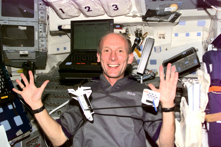 Das Bild zeigt den Astronauten Gerhard Thiele lachend und mit ausgebreiteten Armen während eines Flugs mit dem Space Shuttle. Vor und hinter ihm schweben mehrere Gegenstände, u.a. das Modell eines Space Shuttles.