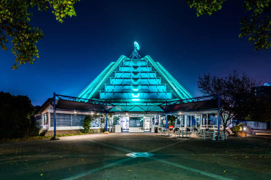 Das Bild zeigt den Eingangsbereich des Planetarium Mannheim bei Nacht aus der Nähe. Die Kuppel ist hellblau erleuchtet. 