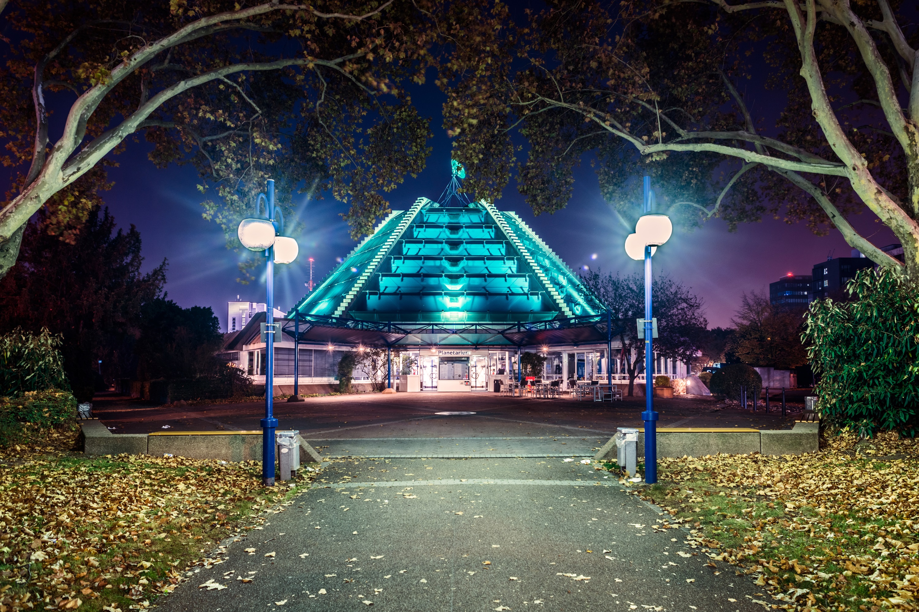 Das Bild zeigt den Eingangsbereich des Planetarium Mannheim bei Nacht aus etwas größerer Entfernung. Die Kuppel ist hellblau erleuchtet. 