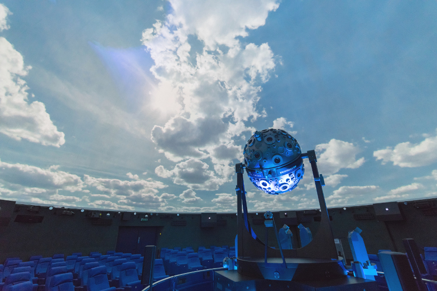 Das Bild zeigt den Kuppelsaal des Planetarium Mannheim. Der Sternenprojektor ist etwas rechts der Mitte zu sehen. An der Kuppel sieht man einen blauen Himmel mit weißen Wolken und die Sonne. 