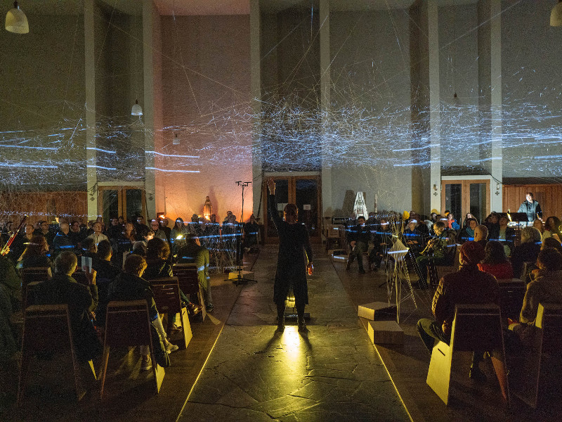 Link zur Beschreibung der Veranstaltung „Atlas Eclipticalis“ am 25.06.2026 - Das Bild zeigt ein Konzert des Ensemble Modern. Menschen sitzen in mehreren Stuhlreihen hintereinander in einem abgedunkelten Saal. In der Mitte steht eine Person.