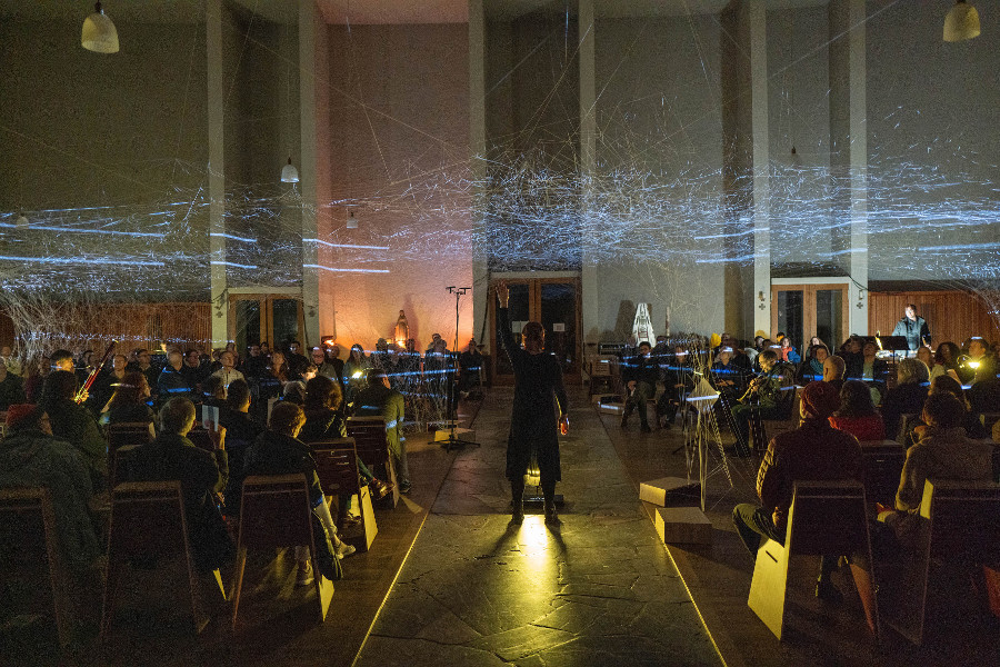 Das Bild zeigt ein Konzert des Ensemble Modern. Menschen sitzen in mehreren Stuhlreihen hintereinander in einem abgedunkelten Saal. In der Mitte steht eine Person. Sie streckt den linken Arm hoch nach oben.
