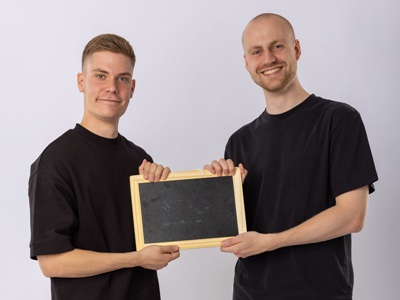 Link zur Beschreibung der Veranstaltung „Mathe kann jeder!“ - Auf dem Bild sind die Vortragenden Johannes Mensing und Josef Naber zu sehen. Sie halten zusammen eine kleine Schultafel zwischen sich.