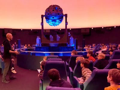 Das Bild zeigt unseren Kuppelsaal mit unserem Sternenprojektor und unseren Vorführer, der mit einer Gruppe Kindern spricht. 