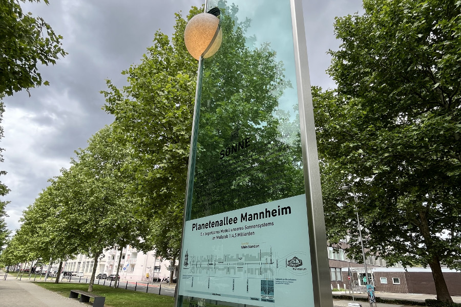 Das Bild zeigt die Sonnenstele der Planetenallee in der Augustaanlage in Mannheim sowie eine Infotafel. Dabei handelt es sich um eine 3,6 m hohe Glasscheibe, an der oben links eine kugelförmige Lampe befestigt ist, die die Sonne darstellt.