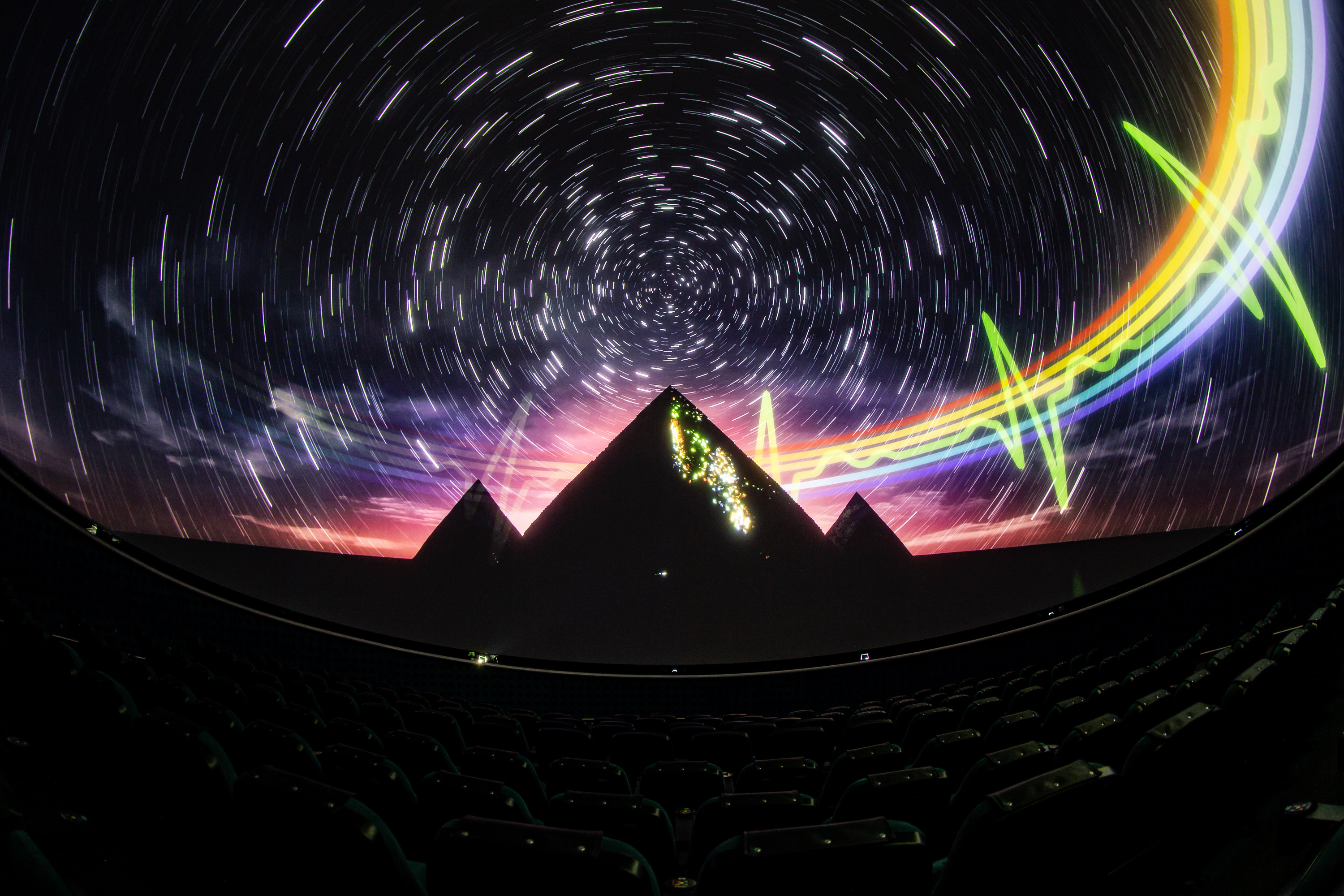 Die künstlerische Darstellung zeigt drei Pyramiden vor dem Nachthimmel. Die Sterne sind zu konzentrischen Strichspuren auseinandergezogen. Ein Regenbogen verläuft im Bildhintergrund. Rechts hat die grüne Linie des Regenbogens die Form eines Herzschlags. Die künstlerische Darstellung zeigt drei Pyramiden vor dem Nachthimmel. Die Sterne sind zu konzentrischen Strichspuren auseinandergezogen. Ein Regenbogen verläuft im Bildhintergrund. Rechts hat die grüne Linie des Regenbogens die Form eines Herzschlags.