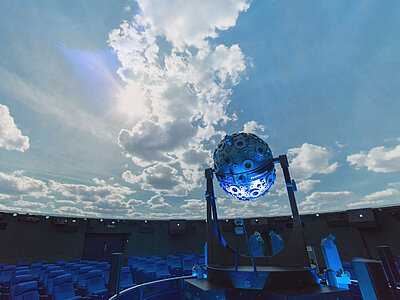 Das Bild zeigt den Kuppelsaal des neuen Mannheimer Planetariums aus dem Jahr 1984. In der Mitte zwischen den konzentrisch angeordneten Sitzreihen steht der kugelförmige Sternenprojektor Universarium vor einer Projektion des Taghimmels an der Kuppel. 