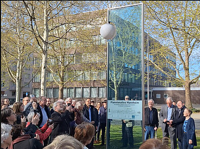 Das Bild zeigt die Sonnenstele der Planetenallee in der Augustaanlage in Mannheim sowie Menschen, die sich um sie versammelt haben, bei der Eröffnung am 8. April 2025.