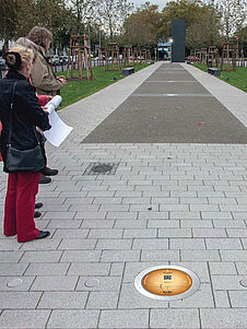 Das Bild zeigt im Vordergrund eine im Boden eingelassene Planetenscheibe mit dem Namen eines Planeten und weiteren Informationen. Im Hintergrund erstreckt sich die Augustaanlage in Richtung Planetarium mit Blick zur Sonnenstele.