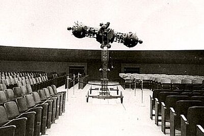 Das Bild zeigt den Kuppelsaal des alten Mannheimer Planetariums aus dem Jahr 1927. In der Mitte zwischen den gerade angeordneten Sitzreihen steht der hantelförmige Sternenprojektor auf einem Rollwagen.