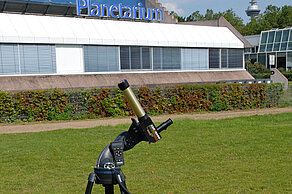 Das Bild zeigt ein Sonnenteleskop, das auf dem Europaplatz hinter dem Planetarium Mannheim steht. Im Bildhintergrund ist ein Teil der Rückseite des Planetariums sichtbar. 