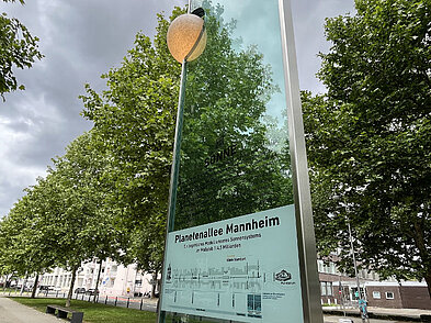 Das Bild zeigt die Sonnenstele der Planetenallee in der Augustaanlage in Mannheim sowie eine Infotafel. Dabei handelt es sich um eine 3,6 m hohe Glasscheibe, an der oben links eine kugelförmige Lampe befestigt ist, die die Sonne darstellt.